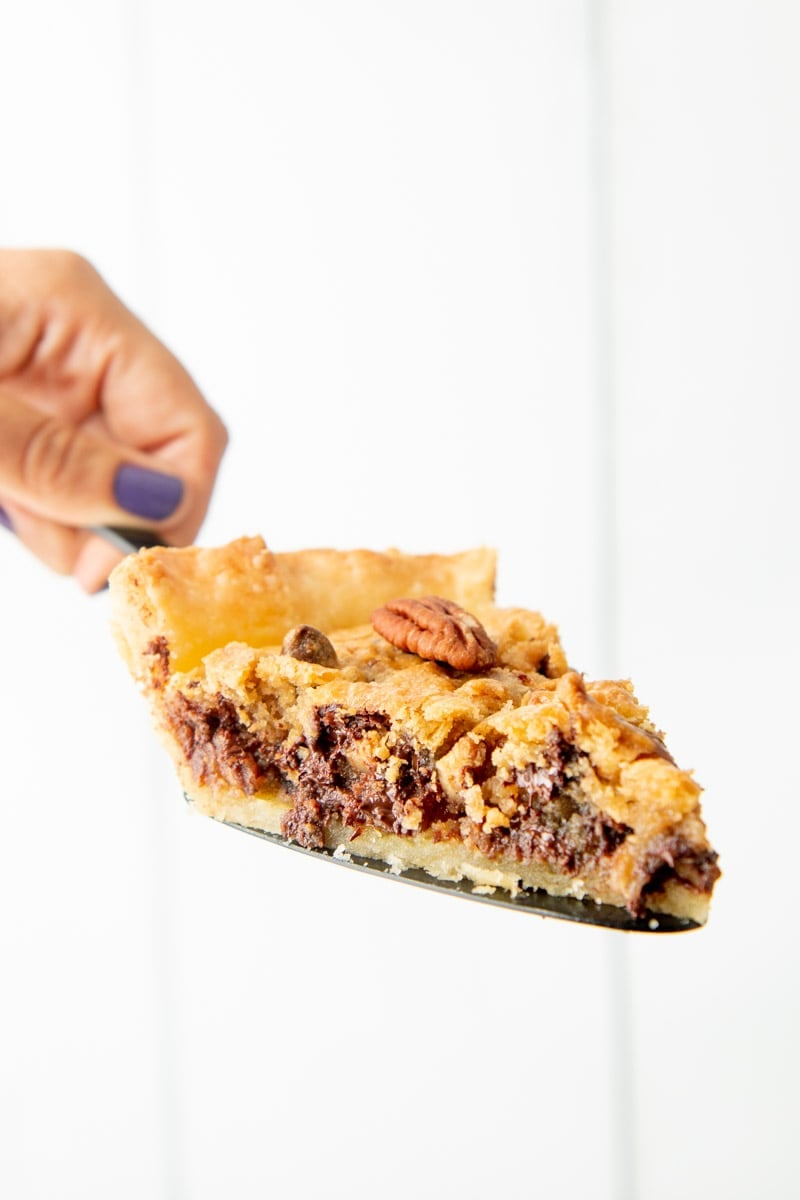 A hand holding a pie server with a slice of chocolate bourbon pecan pie on it.