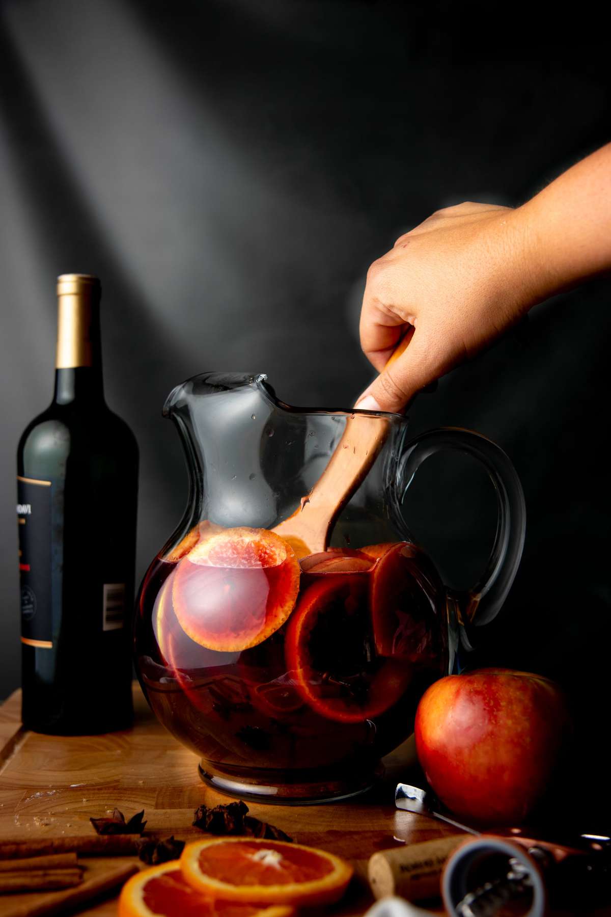 A hand holding a wooden spoon stirs a pitcher with sangria and fruit slices in it.