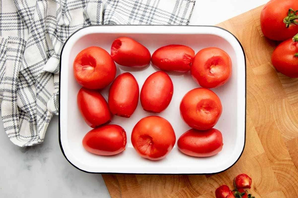Overhead of cored tomatoes in a single layer inside a white baking dish on a wooden cutting board.