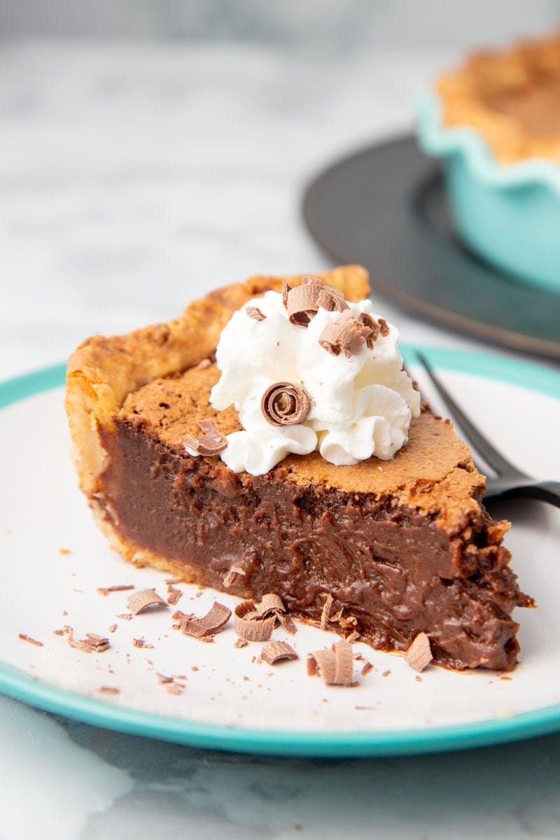 A slice of chocolate chess pie garnished with whipped cream and chocolate curls sits on a plate.
