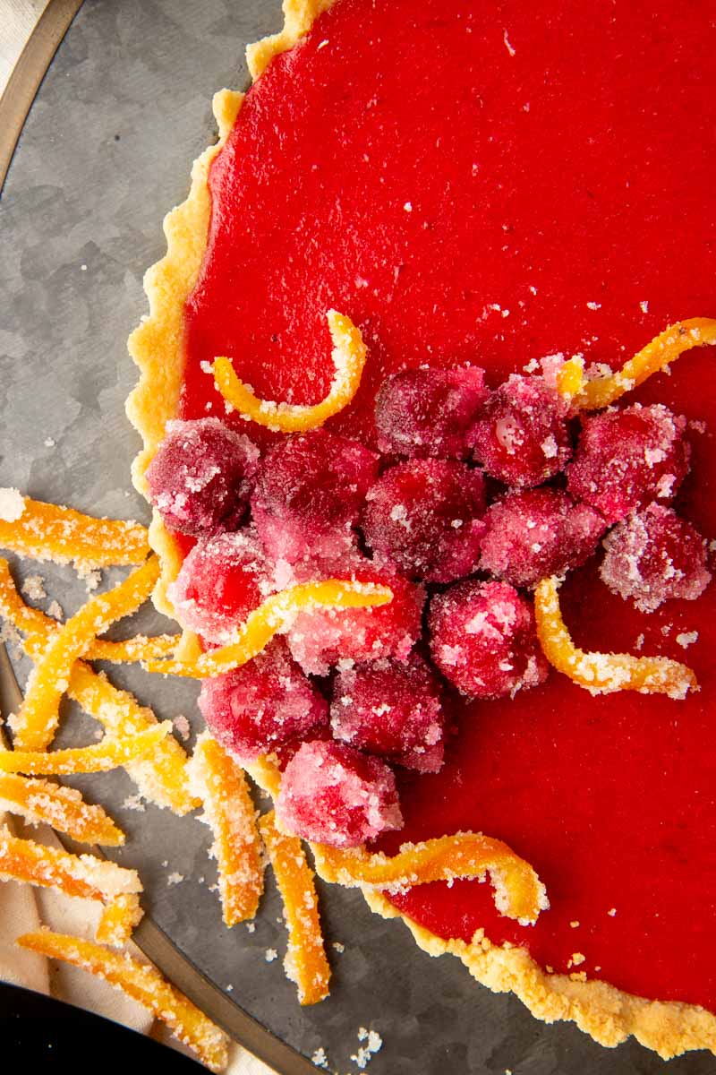 Close view of sugared cranberries and candied orange peel on top of bright red cranberry tart.