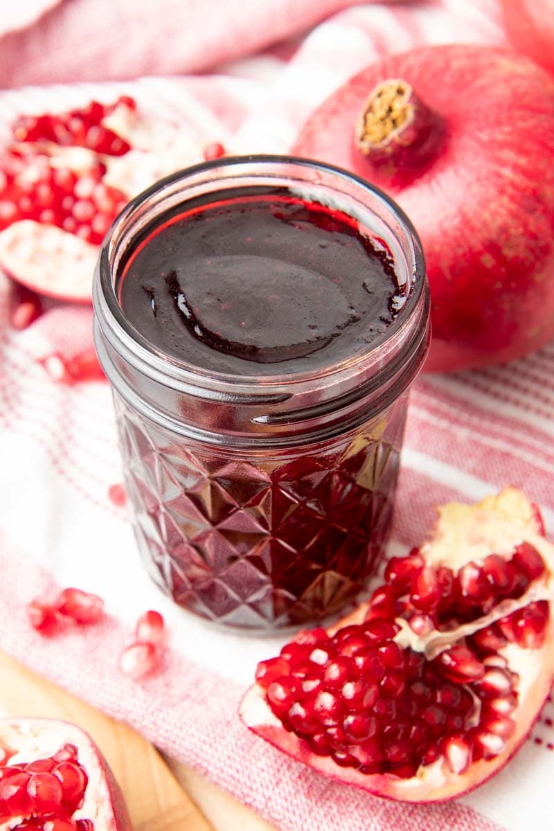 An open jar of pomegranate jelly, perfectly set and surrounded by fresh pomegranates.
