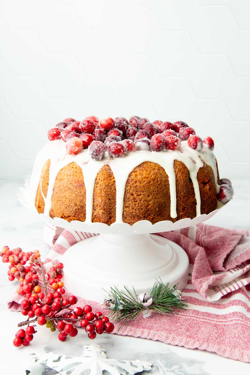 Orange Cranberry Christmas Cake on a white cake stand with sugared cranberries and an orange glaze on top.