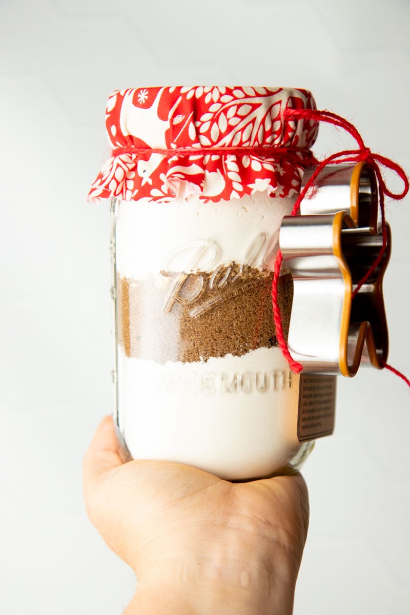 A hand holds up a jar of gingerbread cookie mix packaged for gifting with a cookie cutter.