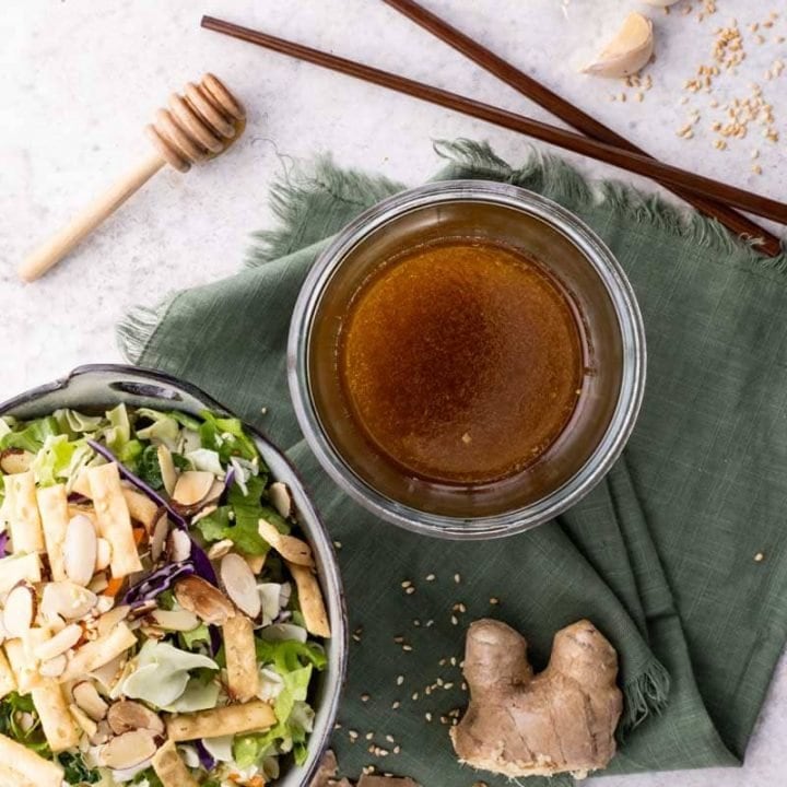 Overhead of sesame ginger dressing in a bowl beside a bowl of salad, fresh ginger, garlic, and sesame seeds around them.