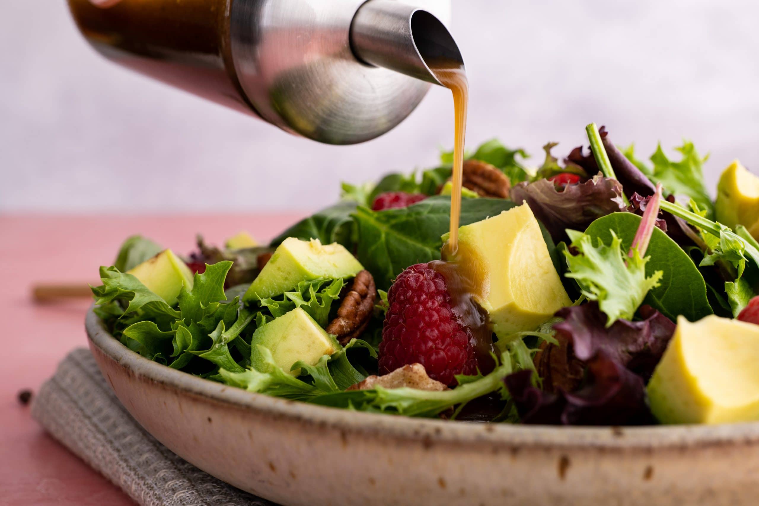 Close up of homemade balsamic vinaigrette pouring onto a fresh green salad topped with chunks of avocado, pecan halves, and raspberries.