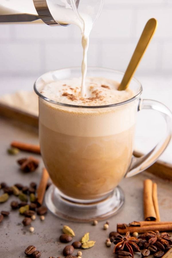 Pouring frothy steamed milk into a glass mug of chai tea with espresso.