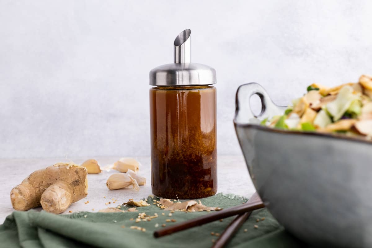 A small glass dressing cruet filled with sesame ginger salad dressing sits beside a big bowl of salad.