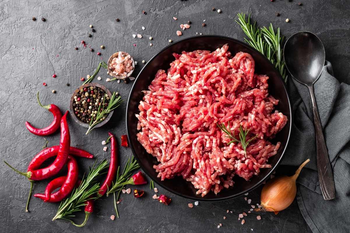 Raw ground beef in a bowl surrounded by seasonings and spices such as salt, pepper, and fresh rosemary.