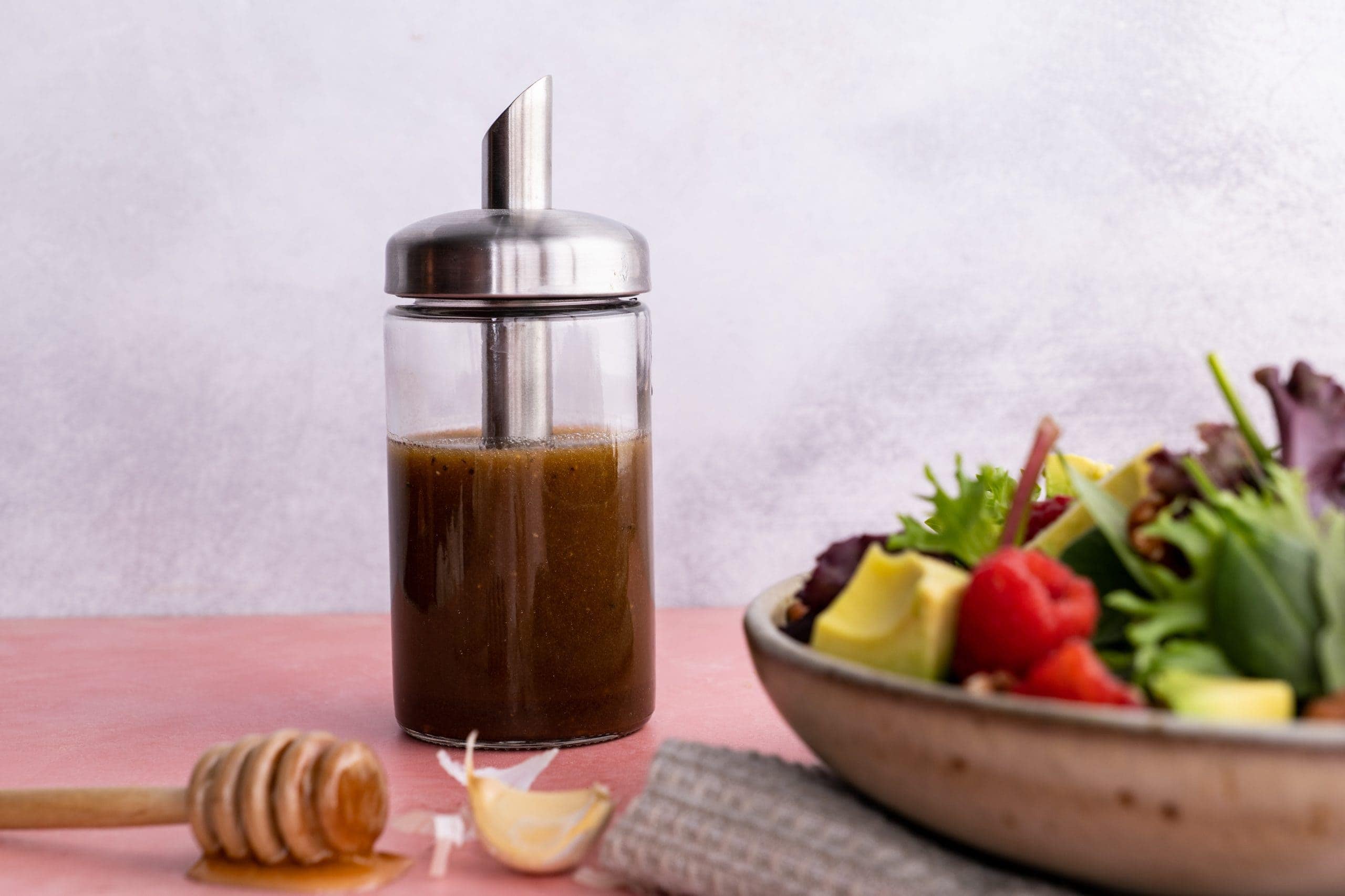 A glass cruet filled with balsamic vinaigrette stands beside a salad bowl filled with fresh ingredients.