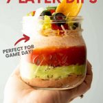 Hand holding a grab and go seven layer dip in a Ball Elite Wide Mouth Pint jar. A text overlay reads, "Grab and Go 7 Layer Dip. Perfect for Game Day! Using Ball Elite Wide Mouth Pint Jars from the Makers of Ball Home Canning Products."