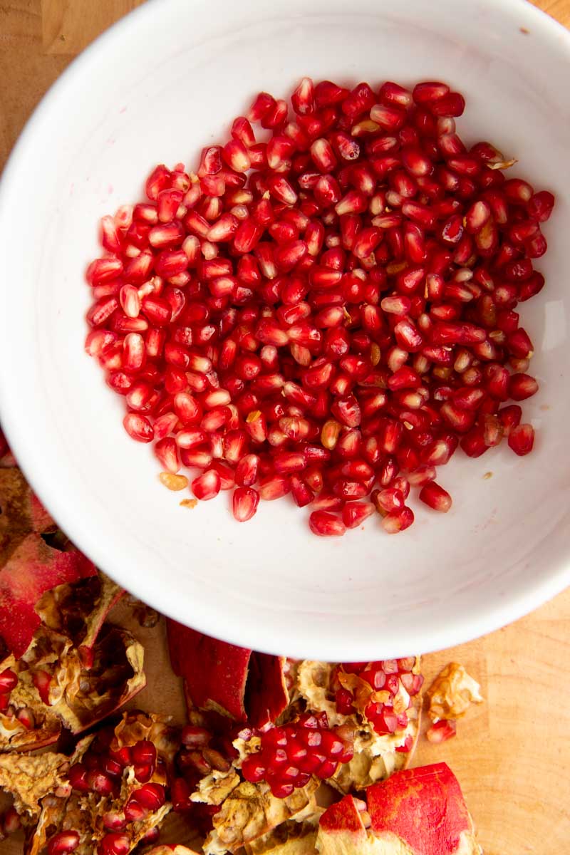 Overhead of pomegranate arils in a white bowl with cut sections of the whole fruit alongside.