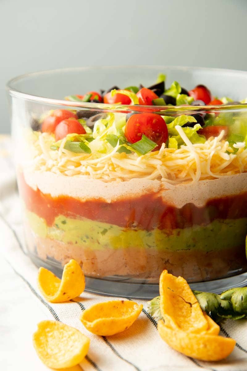 Close view of all seven layers in the taco dip viewed through the tall glass sides of its serving bowl.