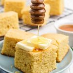 A honey dipper drizzles honey onto a tall square of cornbread topped with two pats of butter.
