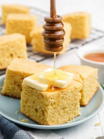 A honey dipper drizzles honey onto a tall square of cornbread topped with two pats of butter.