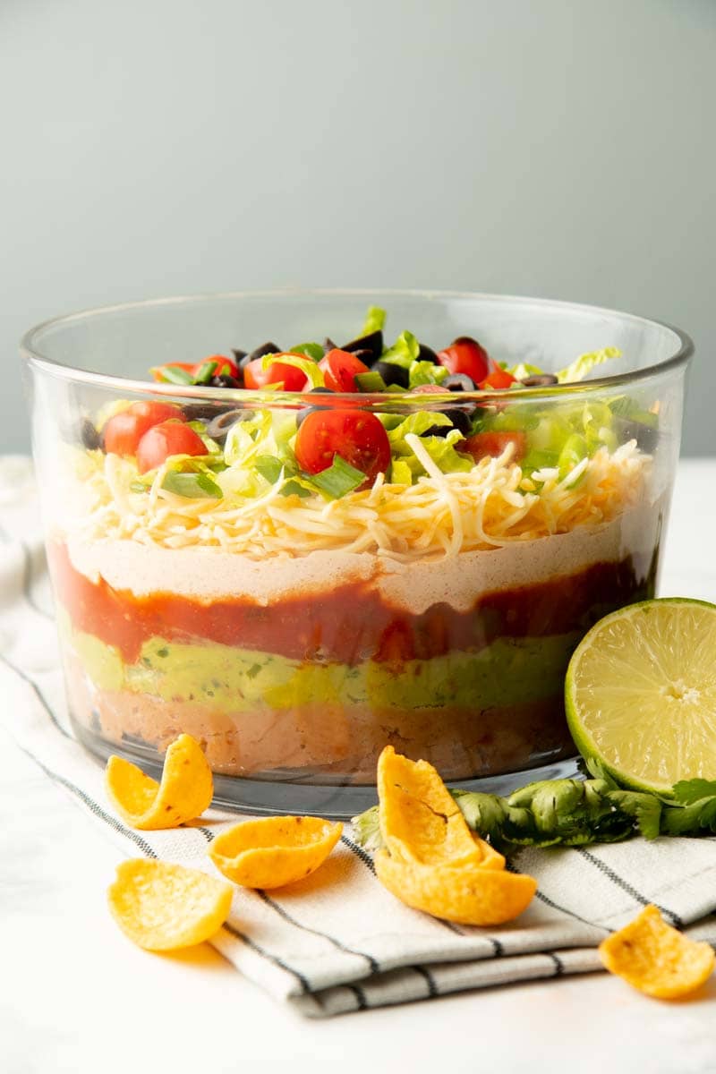Party-sized seven layer dip on a kitchen linen with chips and fresh garnishes around.