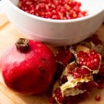 A whole pomegranate and cut sections of fruit stand on a cutting board in front of a bowl of arils.