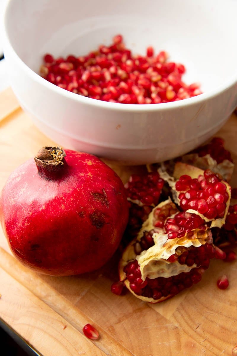 A whole pomegranate and cut sections of fruit stand on a cutting board in front of a bowl of arils.