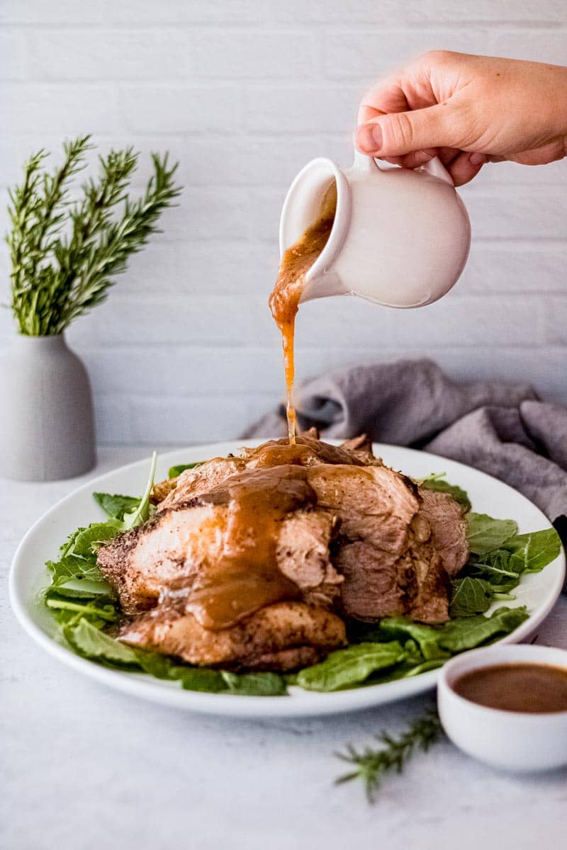 A hand pours gravy down the center of a pile of pork roast slices piled high on a bed of greens.