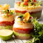 Two individual servings of seven layer dip stand on a kitchen linen in front of a party size bowl of dip.