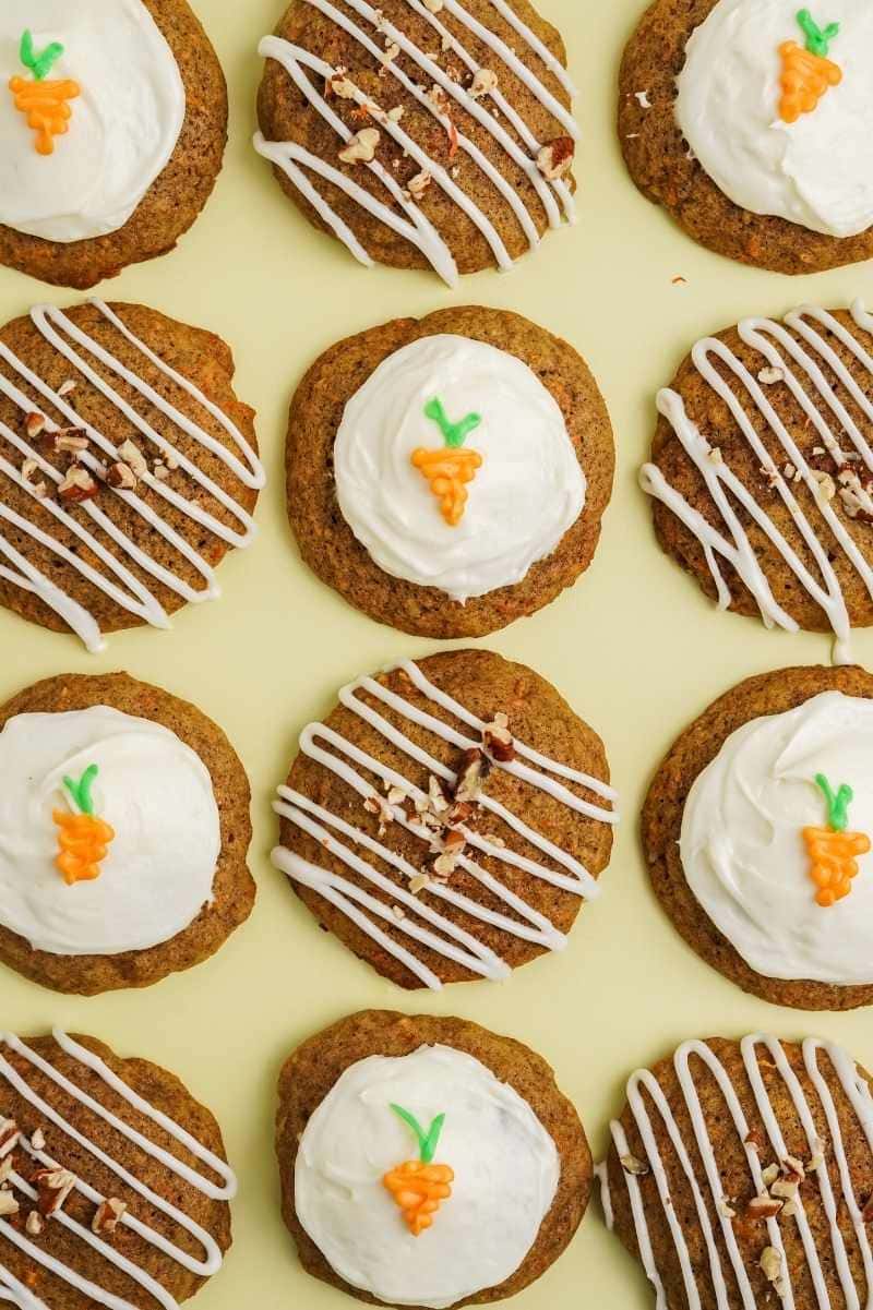 Alternating pattern of soft carrot cake cookies frosted two ways, half with full cream cheese frosting and a frosting carrot detail and half with a frosting drizzle and chopped nuts.