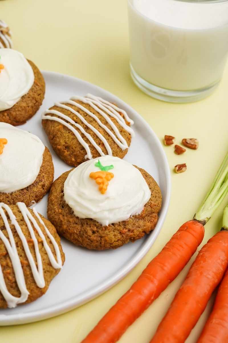 Carrot cake cookies topped with frosting on a white plate alongside a glass of milk.