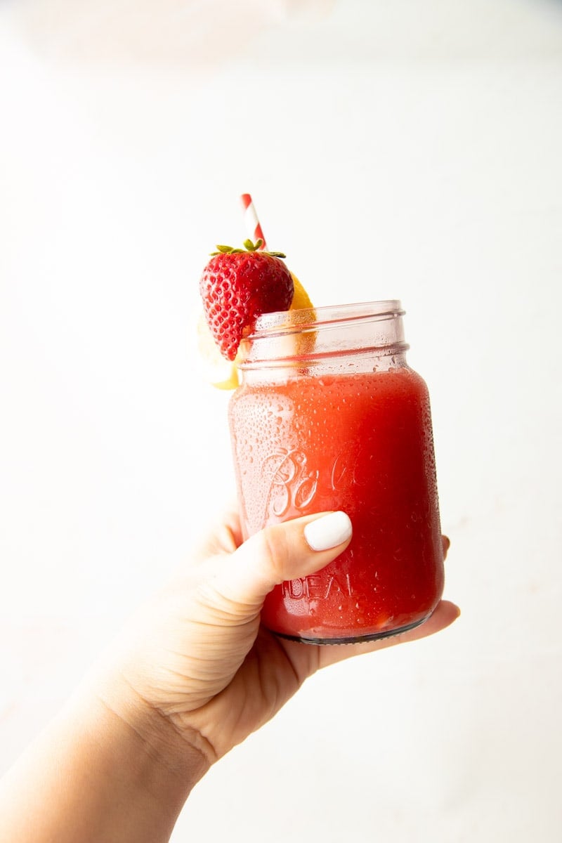 A hand holds out a jar full of strawberry lemonade grnished with a strawberry and slice of lemon. A red and white striped straw sticks out of the drink