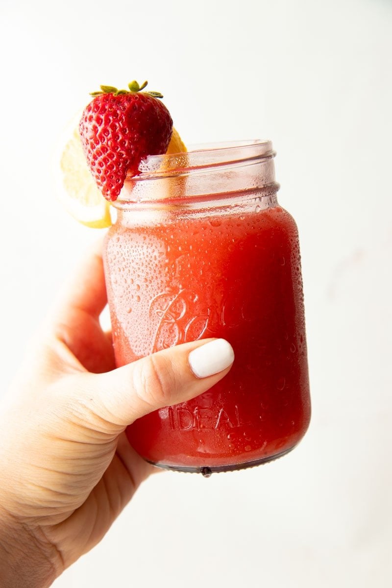 A hand holds out a jar full of strawberry lemonade grnished with a strawberry and slice of lemon