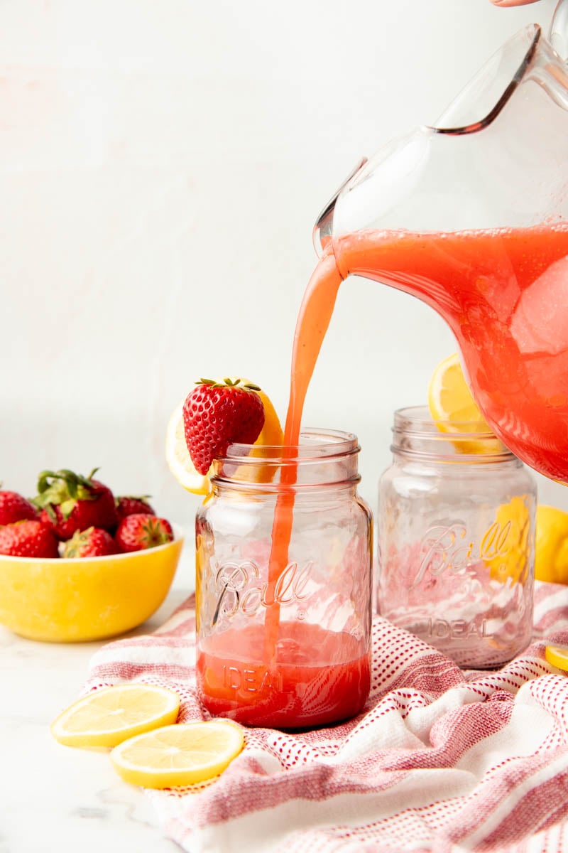 A pitcher pours strawberry lemonade into a pink jar. Strawberries and lemon slices surround the jar