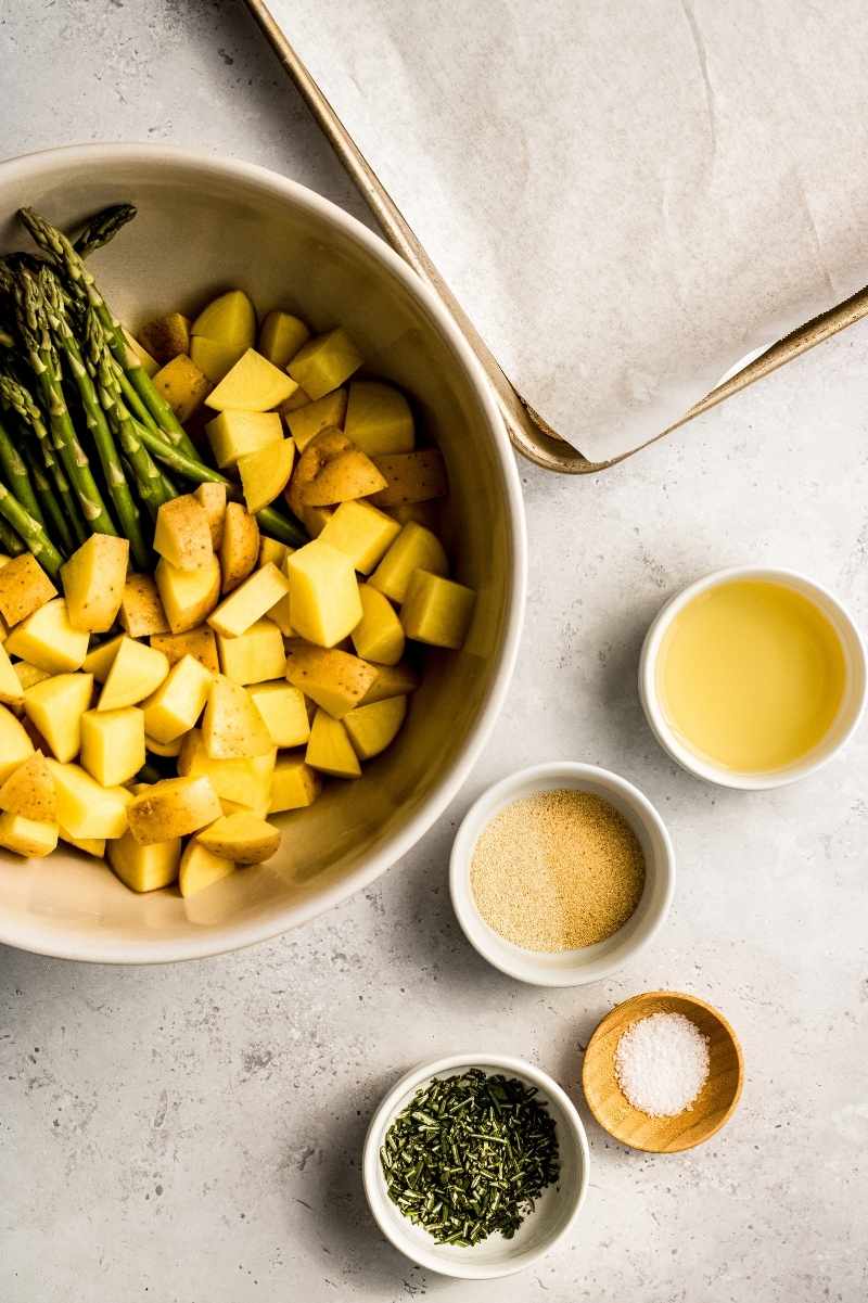 Fresh asparagus and cut potatoes in a bowl with fresh herbs and seasonings alongside.