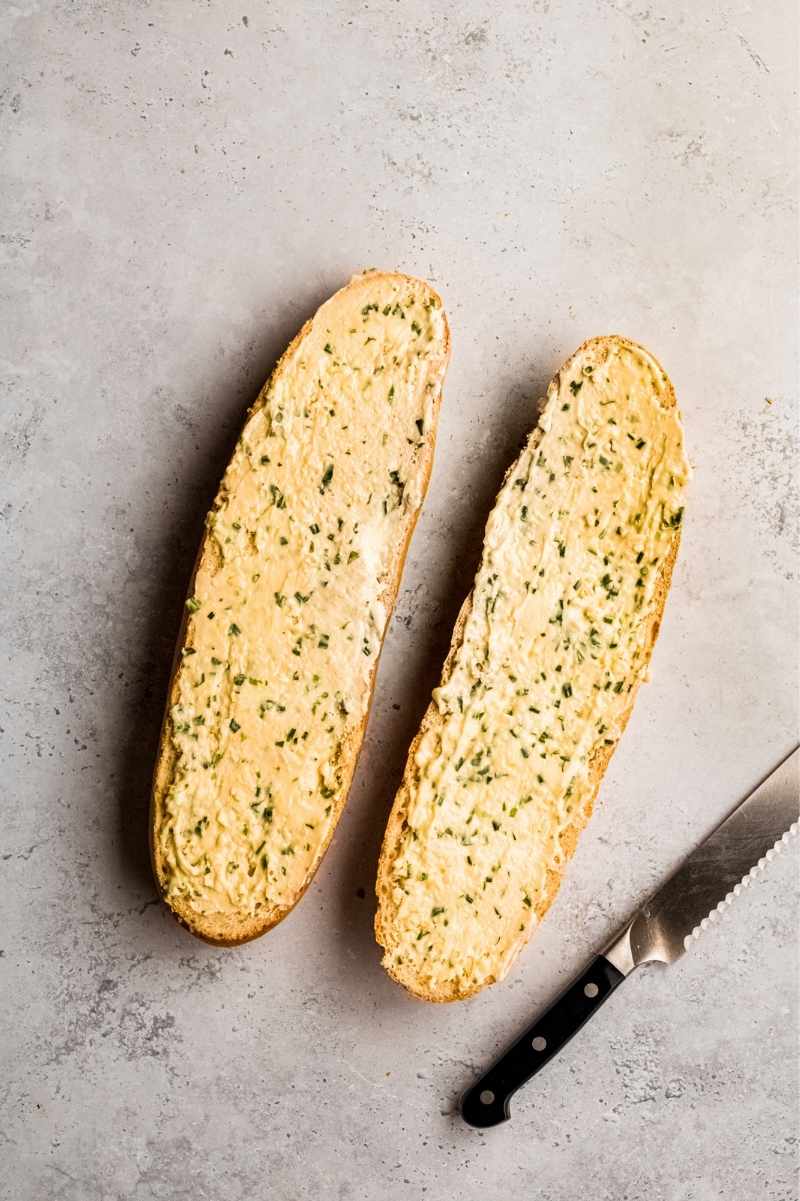 Two halves of a loaf of french bread slathered in homemade garlic butter mixture with a bread knife nearby.
