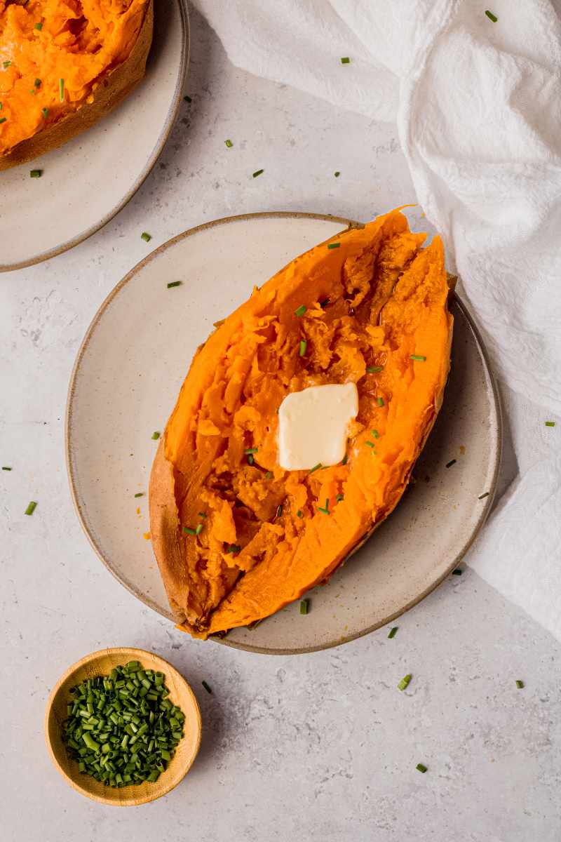 A perfectly cooked sweet potato split open and topped with butter and snipped chives on a plate with a second serving nearby.