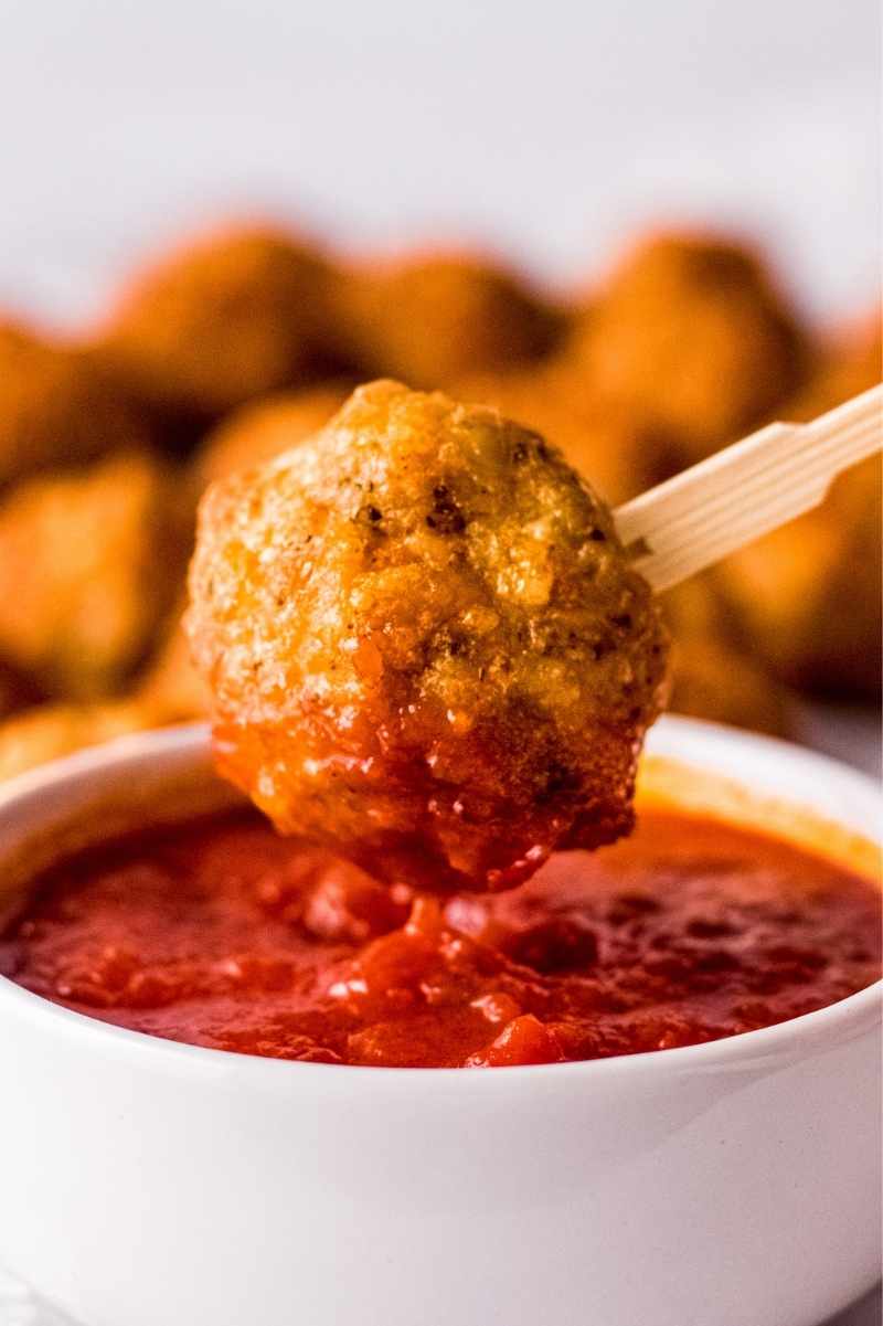 Close view of a baked chicken meatball being dipped into a bowl of marinara sauce on the end of a bamboo cocktail stick.