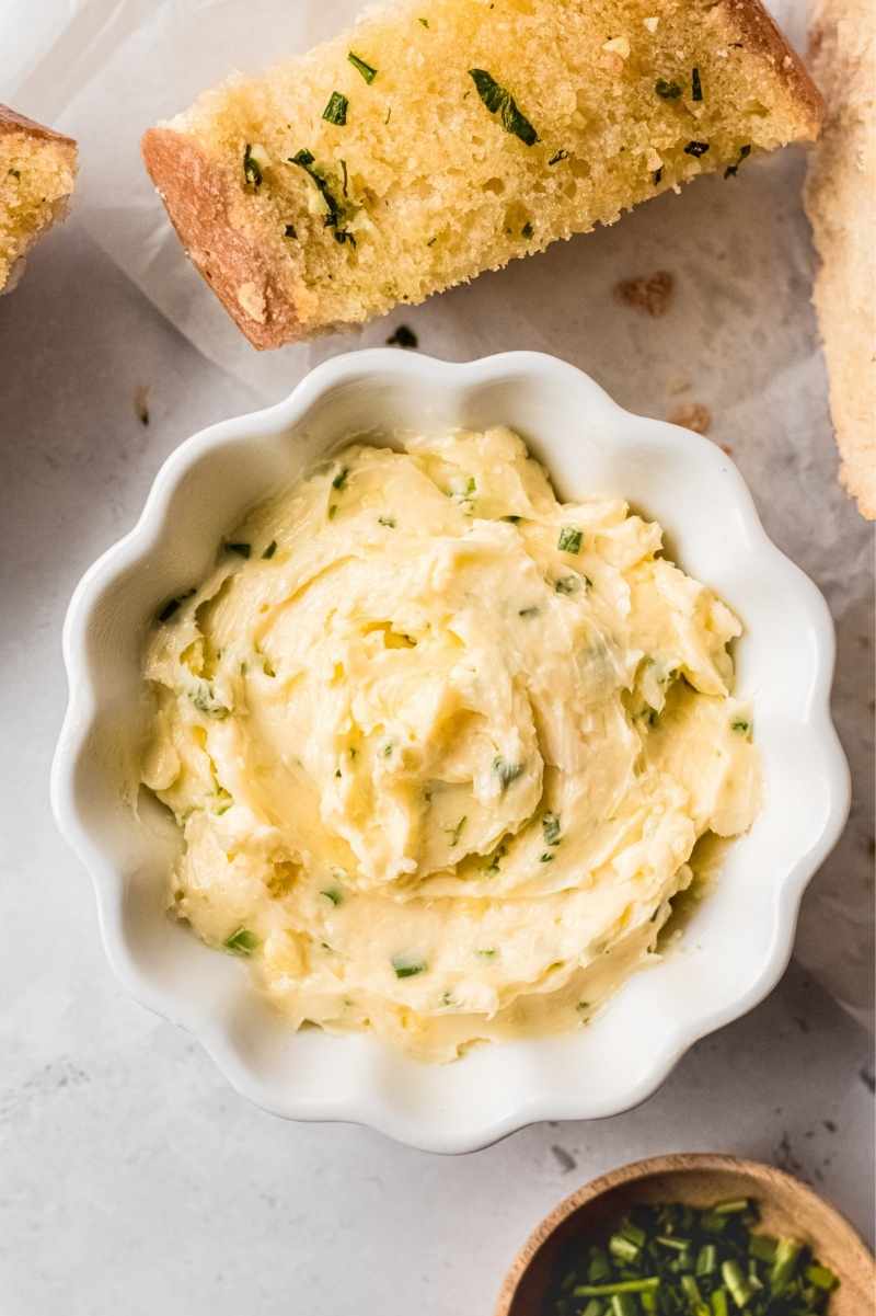 Tight view of garlic butter in a scalloped serving dish with bread and fresh herbs nearby.