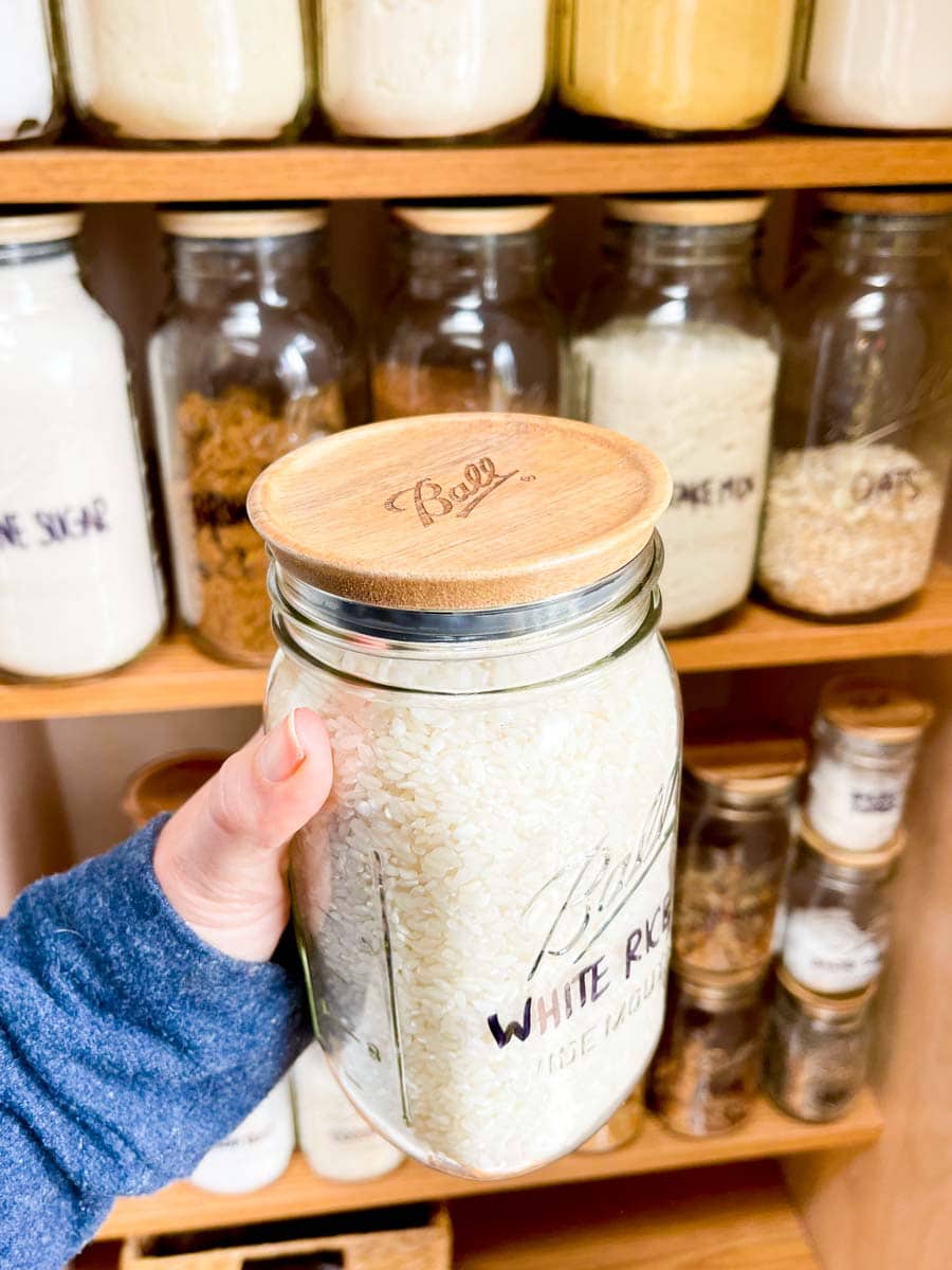 A hand holds a Ball jar filled with white rice capped with an airtight wooden lid in front of an organized pantry.