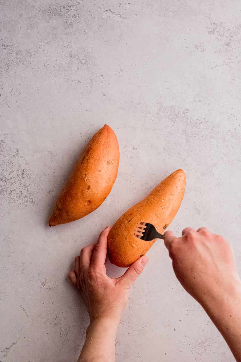Two hands hold a sweet potato and prick the surface with a fork with a second sweet potato nearby.