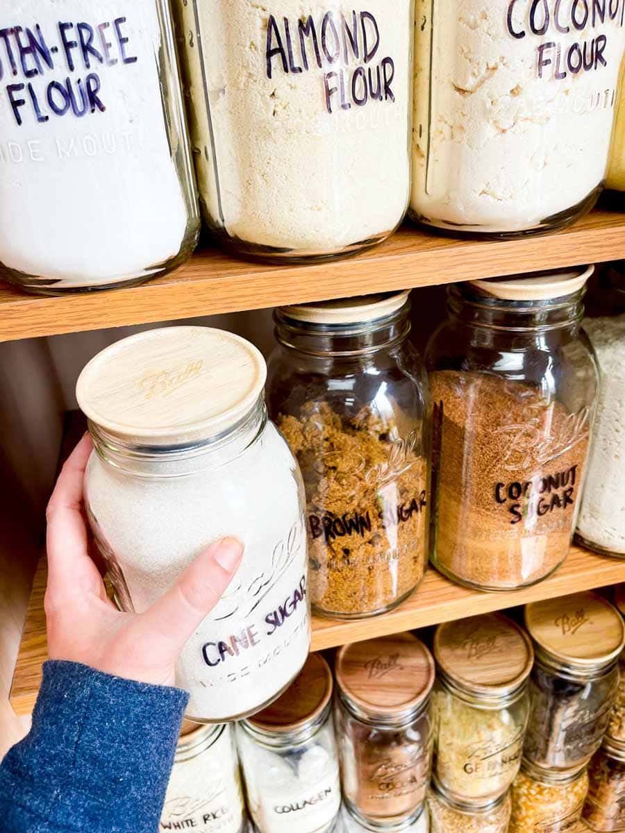 A hand replaces a Ball jar filled with cane sugar on a pantry shelf of baking essentials.