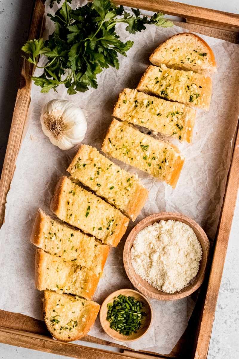 A wooden serving tray lined with parchment paper with sliced garlic bread, fresh parsley, and small bowls of herbs and parmesan cheese on it.