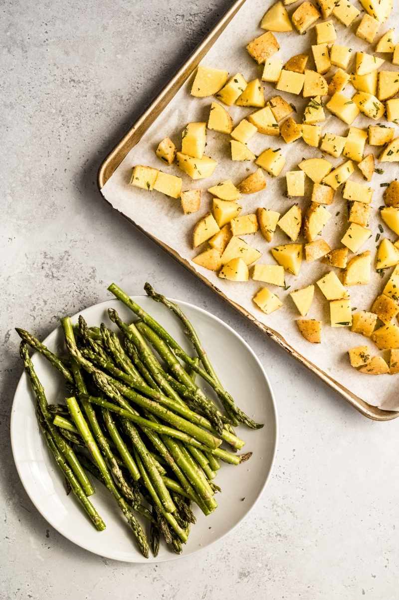 Cut and seasoned potatoes on a sheet tray with seasoned asparagus spears on a plate alongside.