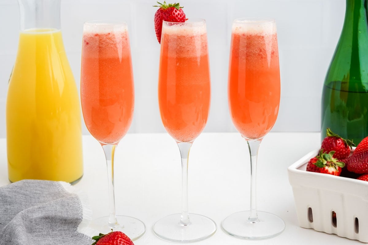 Three stemmed flute glasses filled with pink mimosas stand in a row on a white counter.