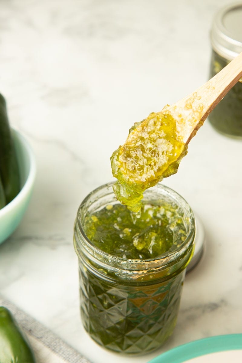 A spoon full of jalapeno pepper jelly hovers above an open quilted pint jar of jelly.