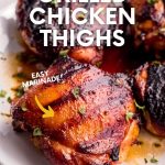 Four juicy grilled chicken thighs on a white plate sitting in flavorful juices.. A text overlay reads, "Grilled Chicken Thighs. Easy Marinade!"