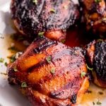 Four juicy grilled chicken thighs on a white plate sitting in flavorful juices..