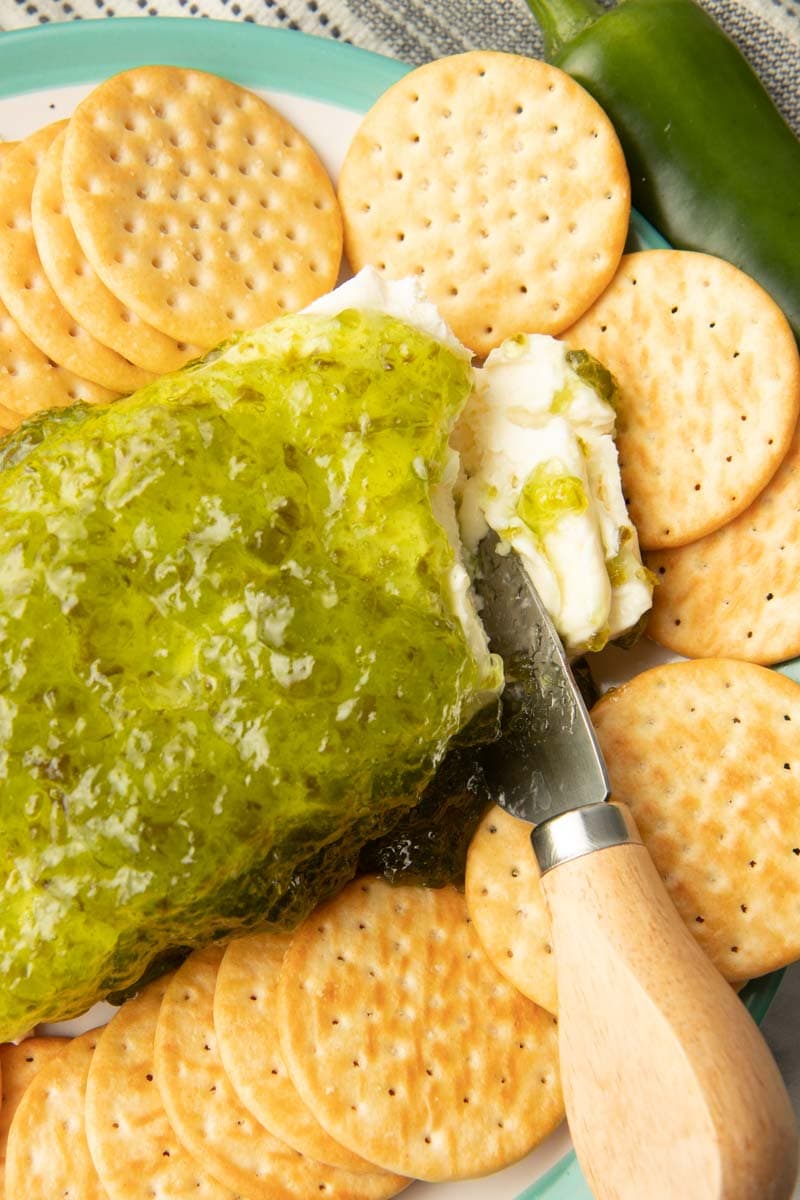 A cheese knife slices into a brick of cream cheese topped with jalapeno pepper jelly on an appetizer platter with crackers.