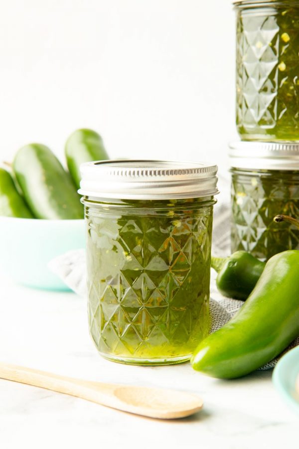 Fresh green jalapeno peppers and a wooden spoon rest beside a jar of pepper jelly.