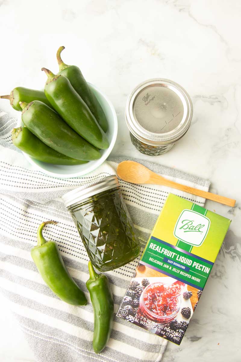 Top view of a jar of jalapeno jelly lying on a tea towel with fresh peppers and a box of liquid pectin beside it.