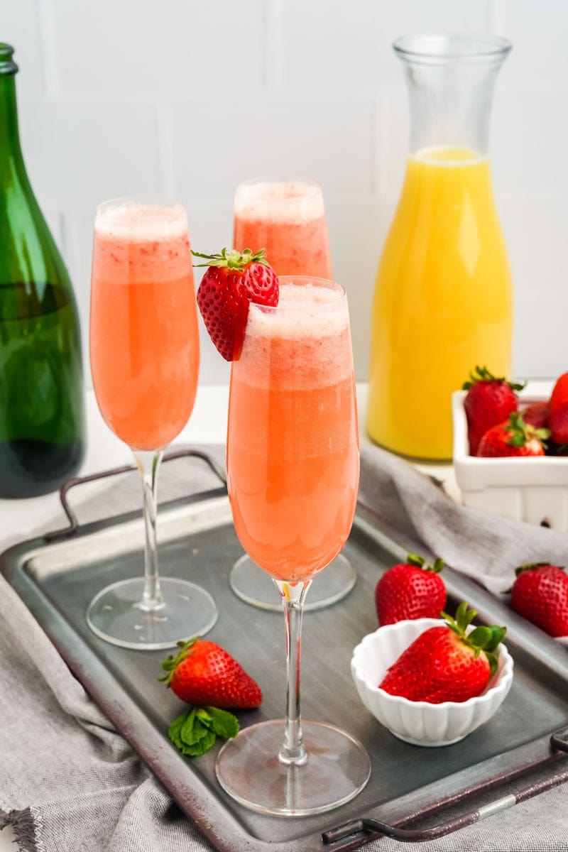 A full champagne flute with a red strawberry on the rim stands on a serving tray with two additional drinks behind it.