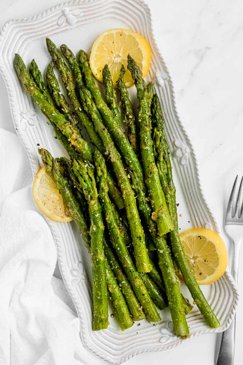 A white, rectangular serving platter with seasoned, steamed spears of asparagus arranged with lemon slices and a fork nearby.