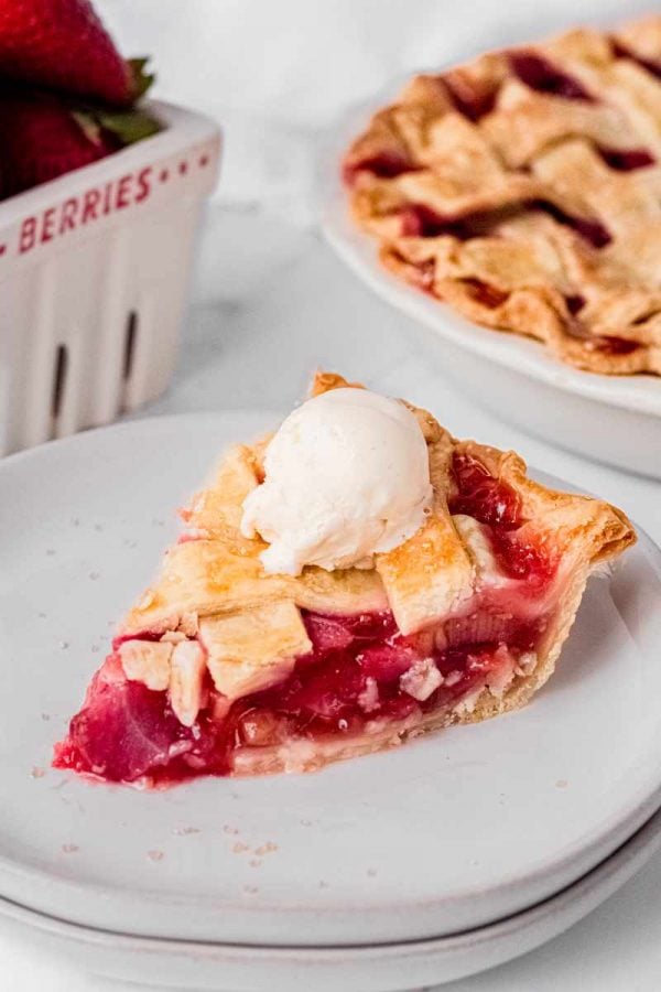 A single slice of strawberry rhubarb pie on a white plate topped with a small scoop of vanilla ice cream.