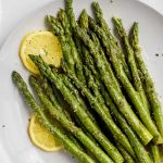 Close view of seasoned, plump asparagus on a round white plate with two lemon wheels.